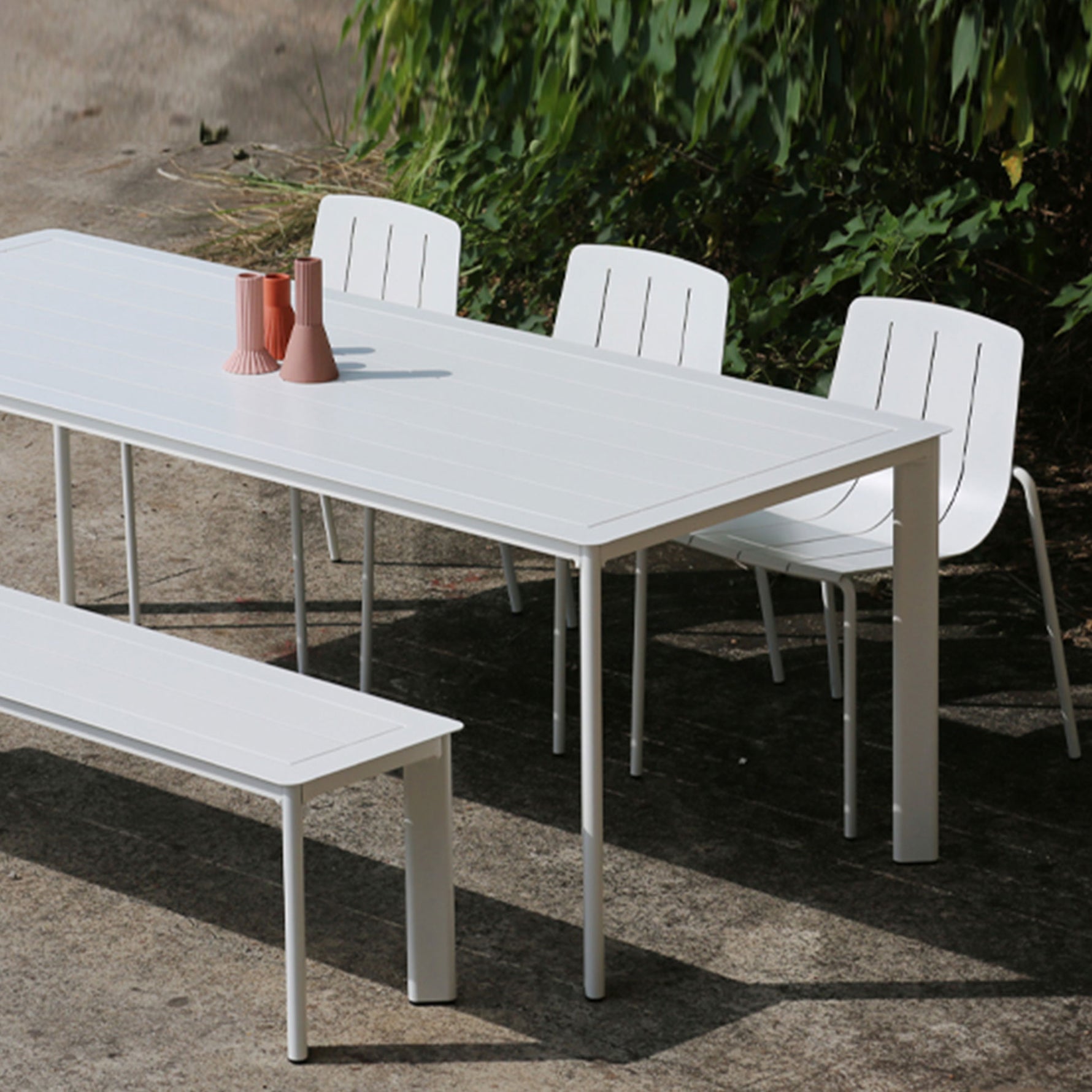 An outdoor dining set comprising of a rectangular dining table, one bench, and four chairs. The set is colored in a light grey finish, and is placed outdoors with some plants in the background.