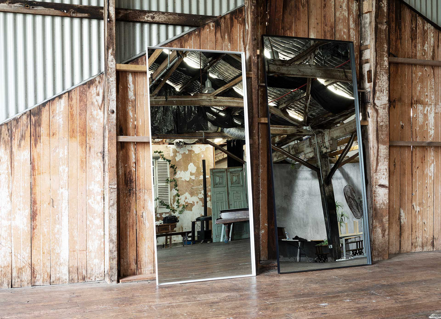 Side-by-side comparison of Luca Mirrors in black and white, showing both standard and oversized sizes in a raw warehouse setting to highlight scale and finish differences
