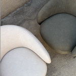 Two modern curved armchairs upholstered in soft textured fabric, shown from above on a concrete floor. One chair is a light warm gray/beige and the other is a muted gray-green.
