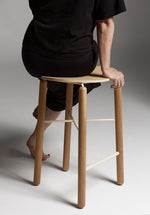 Person seated on the Greta bar stool in oak, showing comfort, seat height, and the natural timber finish in a casual setting