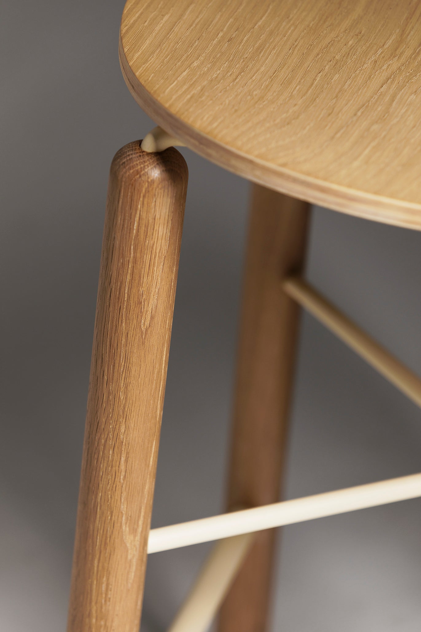 Close-up of the Greta stool in oak showing the curved timber seat and slim black metal leg with footrest, highlighting material detail and construction