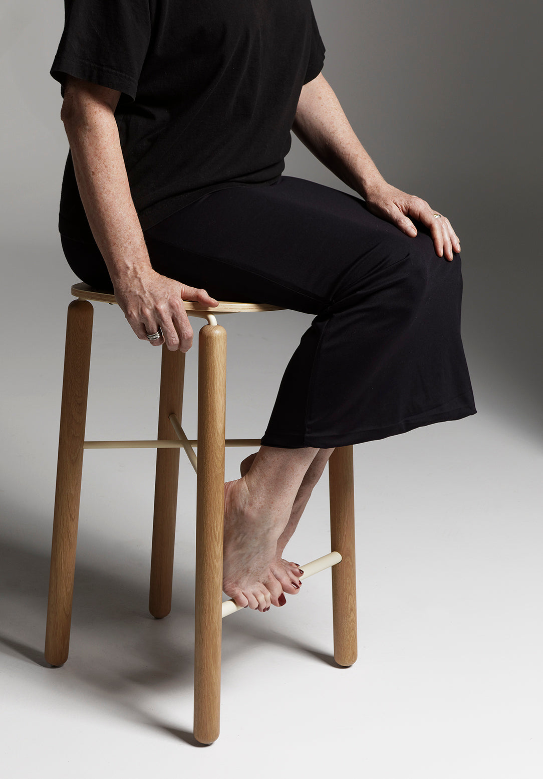 Person casually interacting with the Greta kitchen stool in oak, showing human scale and the stool’s lightweight, minimal design in a studio setting