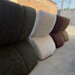 Several textured fabric seat or ottoman cushions in different colors are stacked outdoors on a concrete surface, with a brick building and gate in the background.