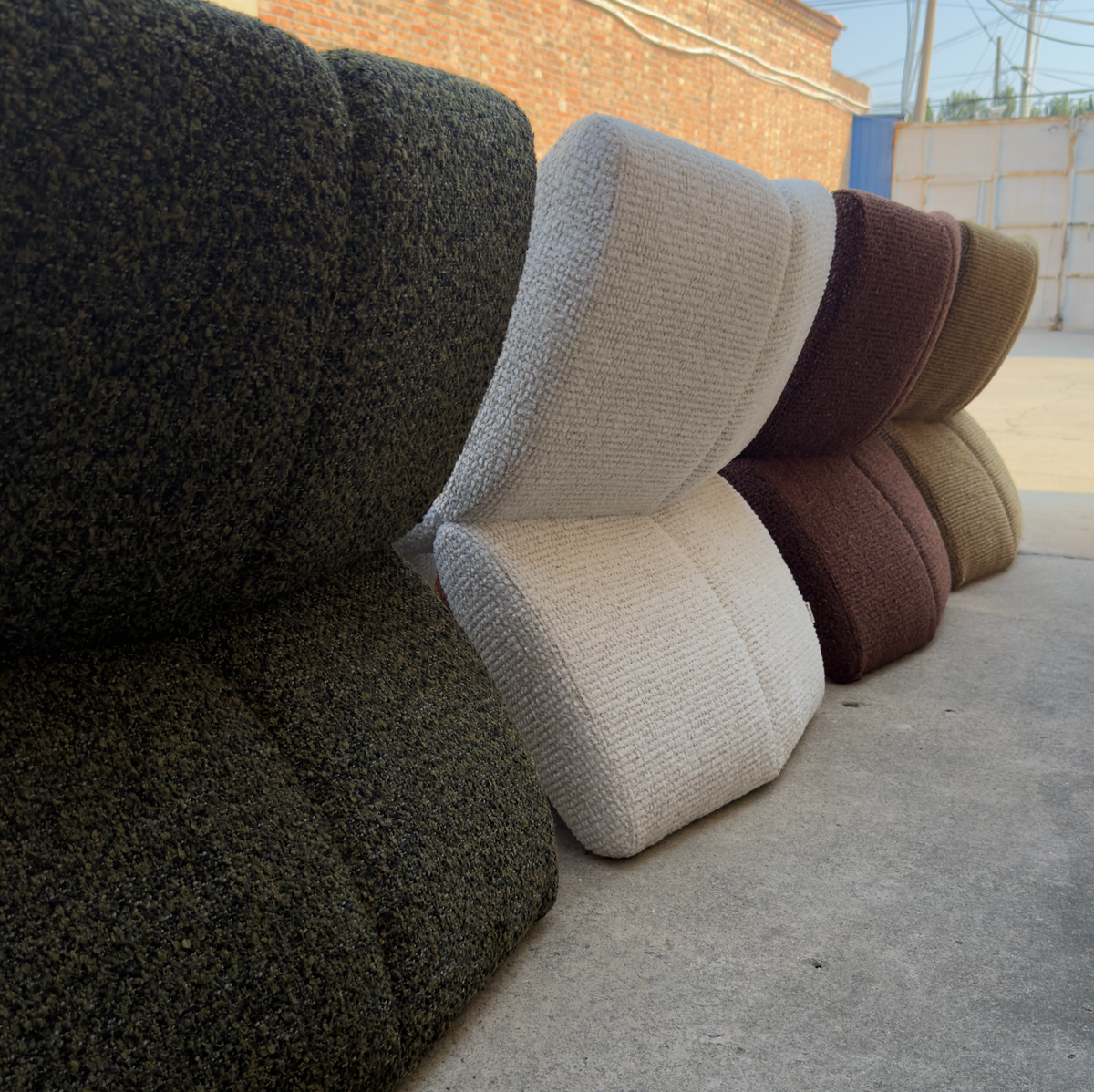 Several textured fabric chair cushions or small upholstered seat modules in multiple colors, including dark green, cream, brown, burgundy, and tan, arranged on a concrete floor outdoors.