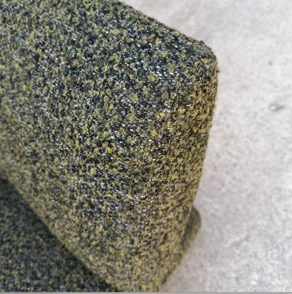 Close-up of a textured fabric or carpet sample with a speckled olive green, black, and gray pattern, shown against a light gray concrete-like background.