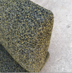 Close-up of a textured fabric or carpet sample with a speckled olive green, black, and gray pattern, shown against a light gray concrete-like background.
