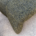 Close-up of a textured woven upholstery fabric in a mottled olive green, black, and tan pattern, draped over the corner of a piece of furniture on a concrete floor.