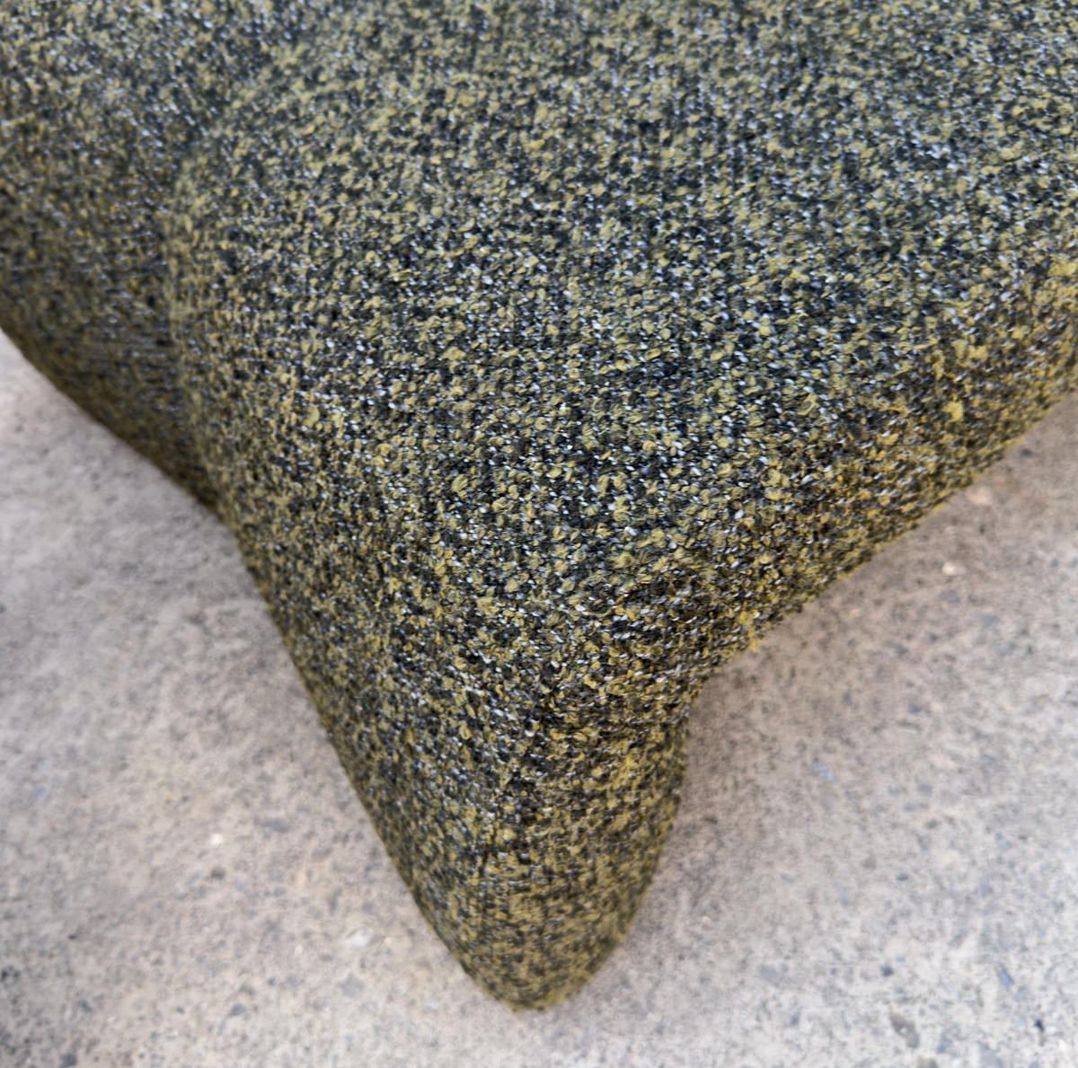 Close-up of a textured woven upholstery fabric in a mottled olive green, black, and tan pattern, draped over the corner of a piece of furniture on a concrete floor.