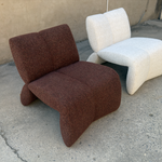 Two modern sculptural lounge chairs with curved tubular forms, one upholstered in dark brown bouclé fabric and the other in off-white bouclé fabric, photographed outdoors on concrete in front of a stucco wall.