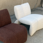 Two modern sculptural lounge chairs with textured bouclé upholstery, one in dark reddish-brown and the other in off-white, photographed outdoors on a concrete floor against a stucco wall.