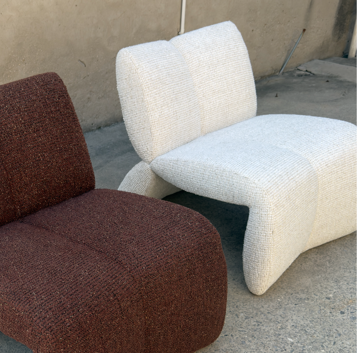 Two modern sculptural lounge chairs with textured bouclé upholstery, one in dark reddish-brown and the other in off-white, photographed outdoors on a concrete floor against a stucco wall.