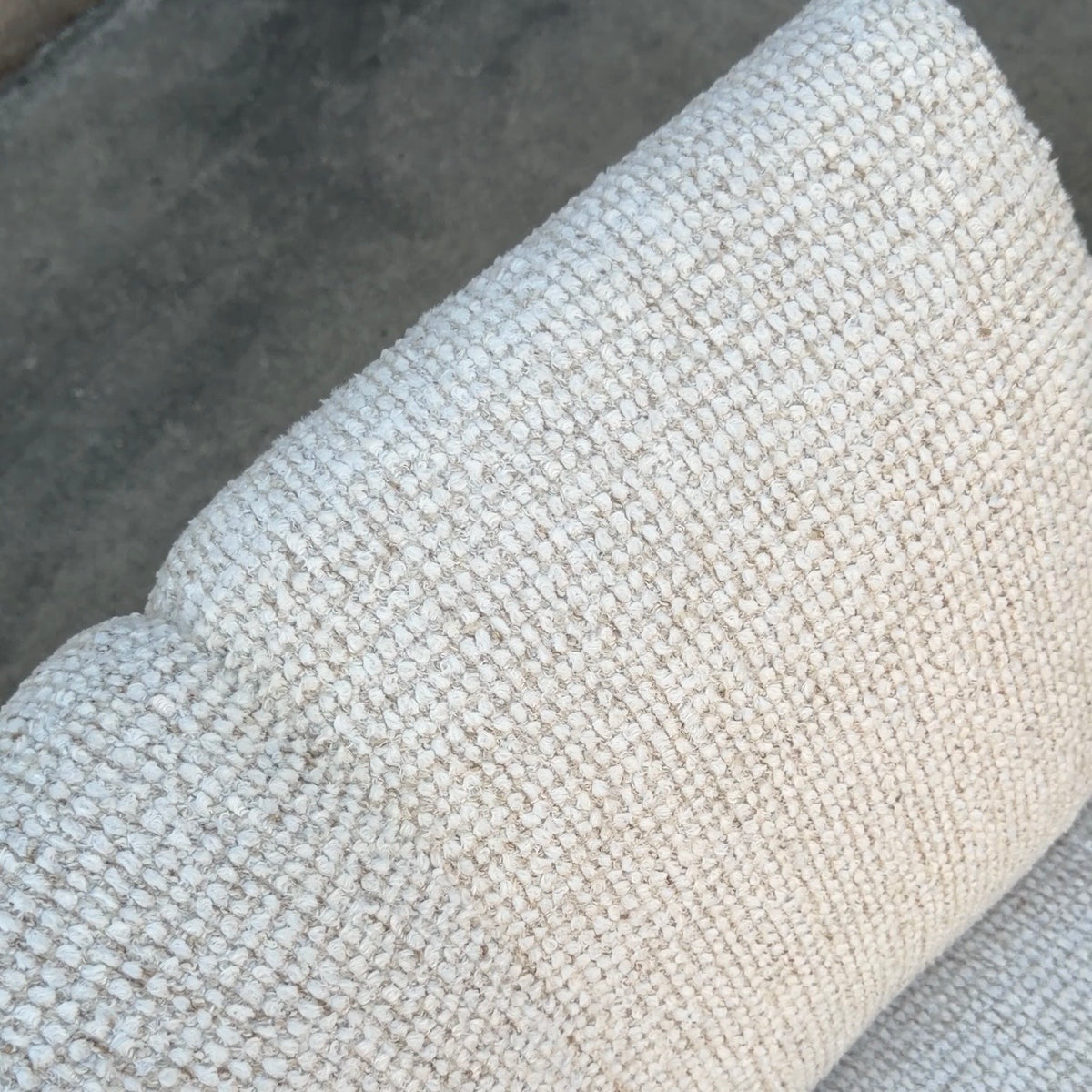Close-up of a textured woven fabric in a warm off-white or cream tone, showing the detailed knit pattern against a darker gray background.