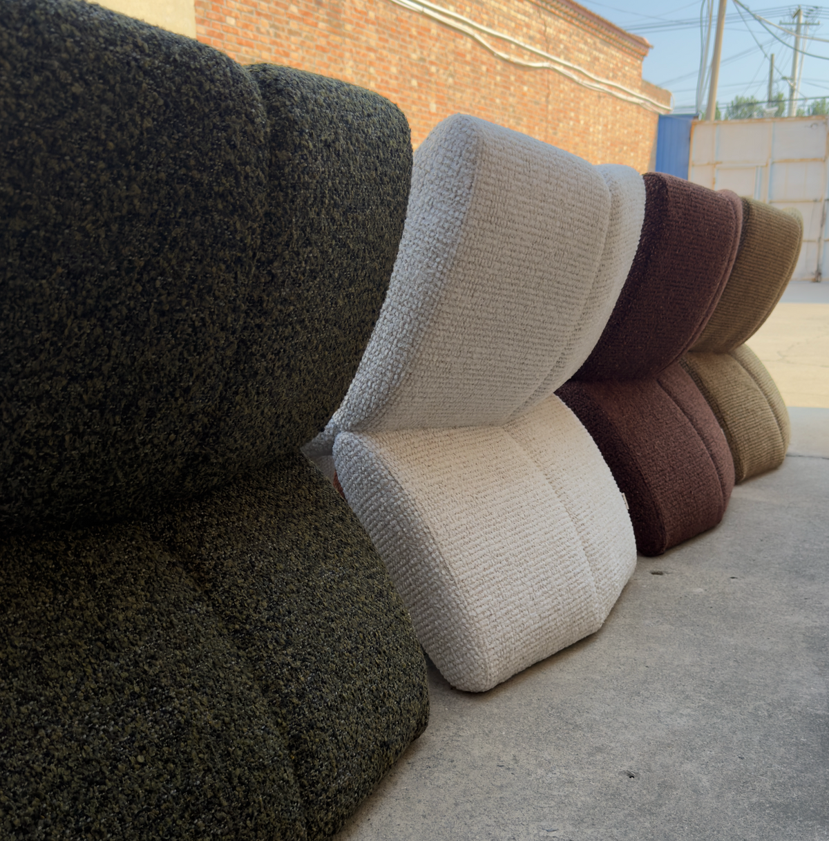 Several textured upholstered chairs in different colors are stacked outdoors on a concrete surface, with a brick building and open yard visible in the background.