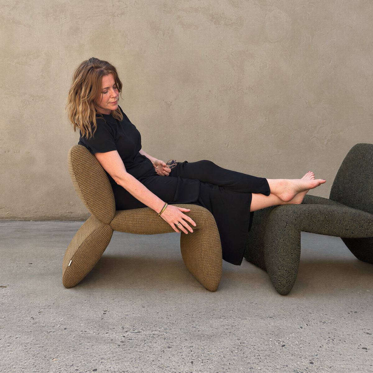 A woman in a black outfit reclines on a sculptural textured armchair in front of a stucco wall, with another dark green chair partially visible behind her.