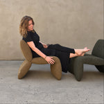 A woman in a black outfit reclines on a sculptural textured armchair in front of a stucco wall, with another dark green chair partially visible behind her.