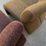 A close-up of a textured woven armchair in warm tan/beige upholstery, with part of a darker reddish-brown chair visible in the foreground on a concrete floor.
