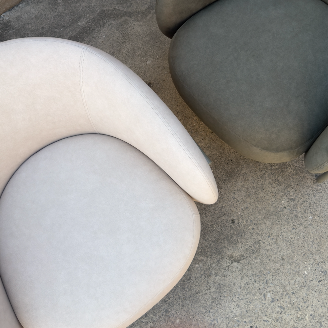 Two modern curved armchairs with rounded seat cushions, one in light beige and one in dark gray, placed on a concrete floor.