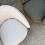 Two modern curved armchairs with rounded seat cushions, one in light beige and one in dark gray, placed on a concrete floor.