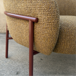 Close-up of a textured woven armchair in warm beige and brown tones with a reddish-brown metal frame, shown outdoors against a concrete wall and ground.