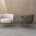 Two modern accent armchairs with curved backs and slim metal legs, set against a textured concrete wall and floor. The chair on the left is upholstered in a light cream fabric, and the chair on the right is upholstered in a muted sage green fabric.