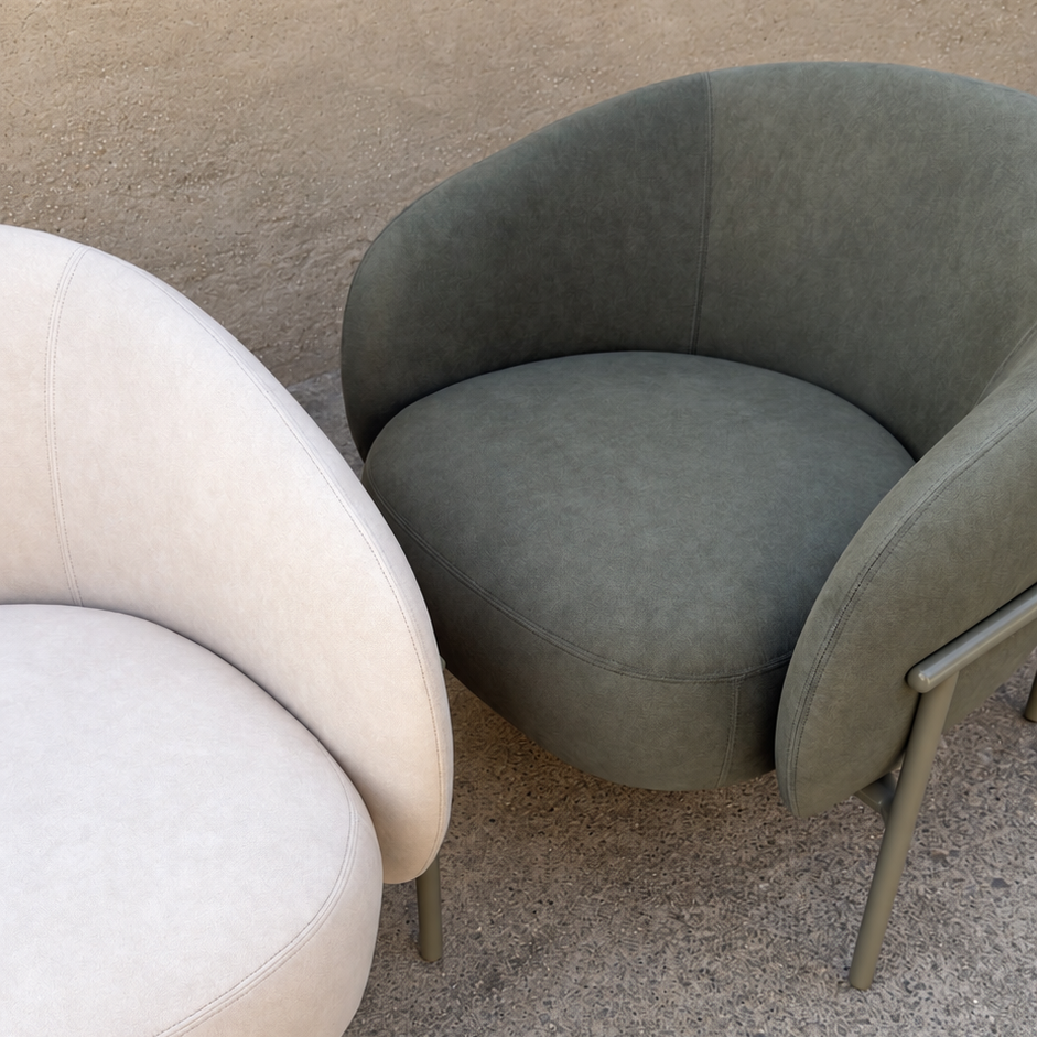 Two modern curved armchairs shown outdoors against a stucco wall and concrete ground. The right chair is upholstered in a muted sage green fabric with slim metal legs, and part of a second chair in a light cream/beige fabric is visible on the left.