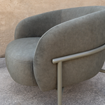A modern curved lounge chair upholstered in textured taupe fabric with a light gray metal frame, set against a neutral beige stucco wall and concrete floor.