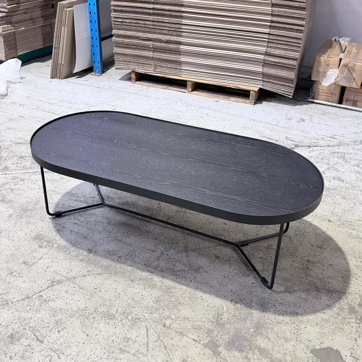 Oval-shaped black coffee table in a warehouse setting with wooden pallets and stacks in the background.