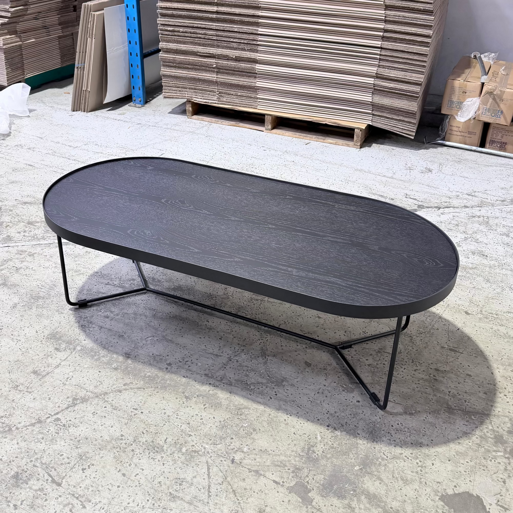 Oval-shaped black coffee table in a warehouse setting with wooden pallets and stacks in the background.