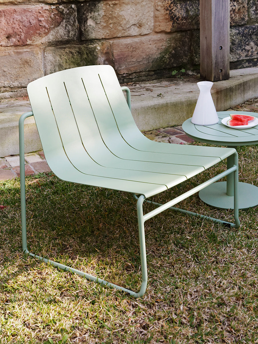 A green outdoor lounge chair with a low-slung profile and sculptural curves, set on grass with a stone wall and side table in the background.