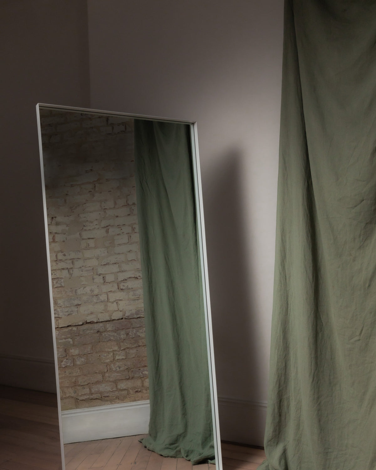 Luca Standard Mirror in white leaning beside a sage linen curtain, reflecting a brick wall