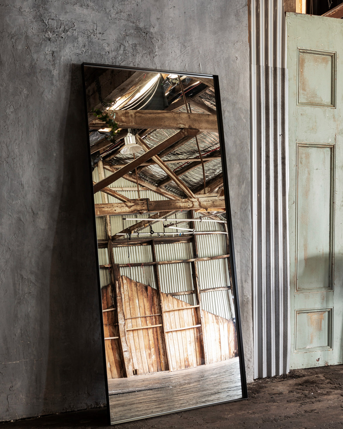 Luca Standard Mirror in black leaning on a concrete wall, reflecting timber rafters